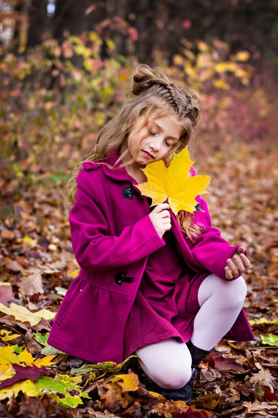 Belle petite fille blonde mignonne dans un manteau rose se tient près du mur avec des feuilles rouges d'automne. — Photo