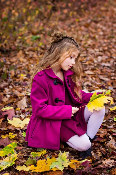 Όμορφη μικρή χαριτωμένη ξανθιά κοπέλα σε ένα ροζ παλτό στέκεται κοντά στον τοίχο με φθινοπωρινά κόκκινα φύλλα. — Φωτογραφία Αρχείου