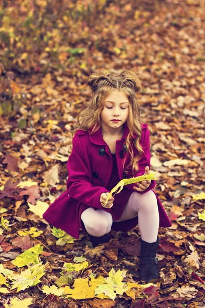 Belle petite fille blonde mignonne dans un manteau rose se tient près du mur avec des feuilles rouges d'automne. — Photo