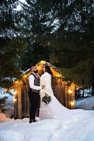 Yeni evli çift, gelin ve damat eski ahşap bir evin arka planında ve eski bir çelenk. Kırsal kış düğünü. Geceleyin düğün töreni. Alışılmadık dekorasyonlar, mumlar, akşam lambaları..