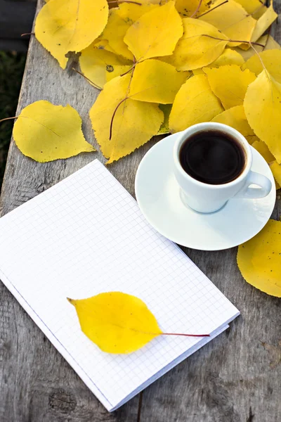 Notebook, cup of coffee and yellow autumn leaves on the old wooden table. — Stock Photo, Image