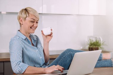 Neşeli genç bir kadın evde otururken hem kahve içiyor hem de bilgisayar başında video konferansında konuşuyor.