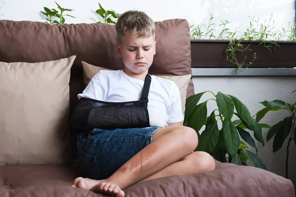 Sad boy with a broken arm and wounds on his leg. Gypsum on a childs hand. — Stock Photo, Image