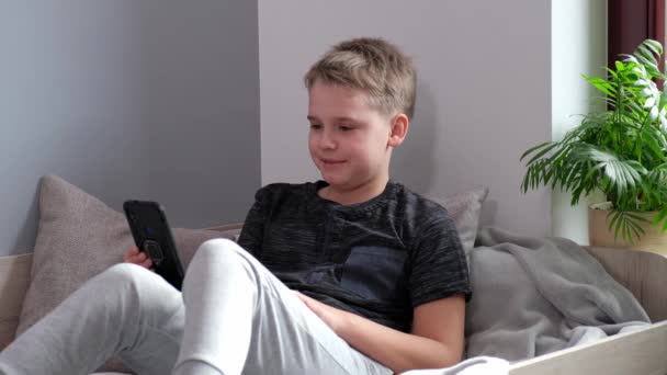 4К Позитивный мальчик улыбается и разговаривает по видео-вызову сидя на кровати в своей комнате. — стоковое видео