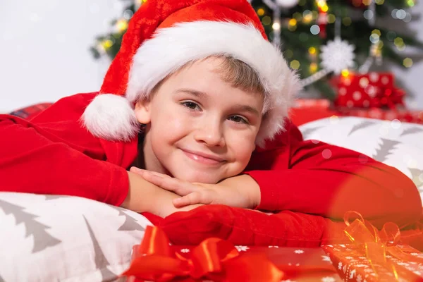 Vacaciones de Navidad. Retrato del niño sonriente en Santa Sombrero Rojo mirando a la cámara, yace rodeado de regalos de Navidad. — Foto de Stock