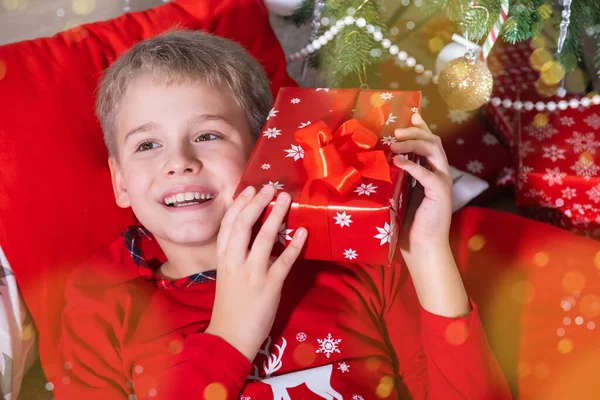 Niño rubio sonriente sosteniendo una caja de regalo festiva y acostado en el suelo cerca del árbol de Navidad — Foto de Stock