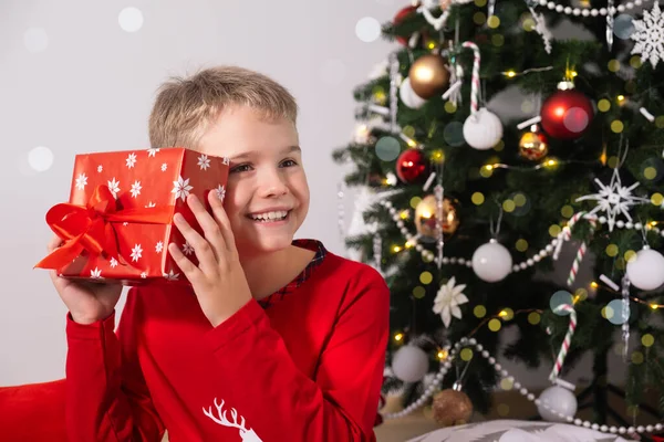 Niño feliz con el pelo rubio está sentado en el suelo en casa y la celebración de regalo de Navidad cerca del árbol de Navidad decorado — Foto de Stock