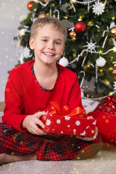Щаслива дитина з світлим волоссям сидить на підлозі вдома і тримає різдвяний подарунок біля прикрашеної ялинки — стокове фото