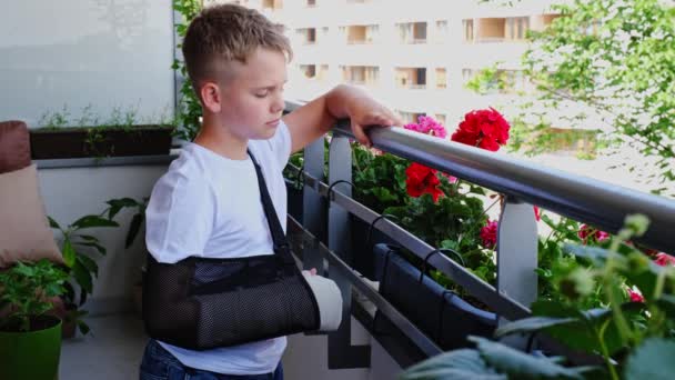 Грустный ребенок со сломанной рукой в гипсе стоит на балконе и машет кому-то рукой — стоковое видео