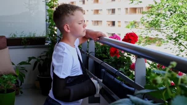 Triest kind met een gebroken arm in een gips kijkt uit in de tuin vanaf het balkon — Stockvideo