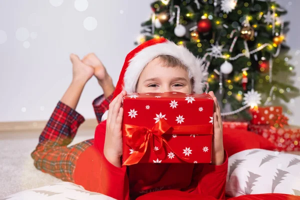 Щасливий милий блондин хлопець у Санта-Капелюсі тримає святкову подарункову коробку і лежить на підлозі біля ялинки — стокове фото