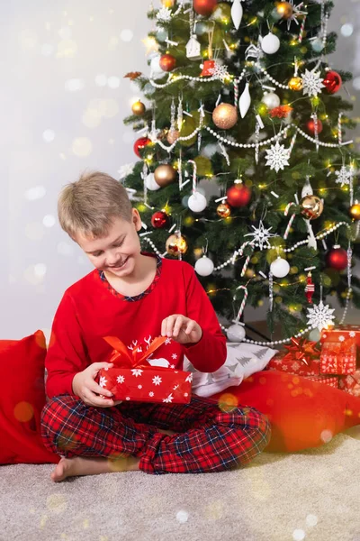 Щасливий хлопчик сидить вдома на підлозі і розбирає різдвяний подарунок біля прикрашеної ялинки. — стокове фото