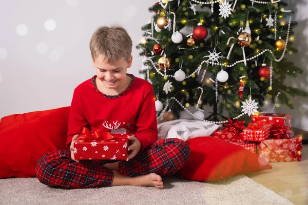 Happy child with blonde hair is sitting on floor at home and holding Christmas present near decorated Christmas tree