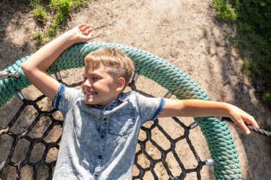 Neşeli sarışın çocuk gülümsüyor ve halka açık bir parkta salıncakta sallanıyor..