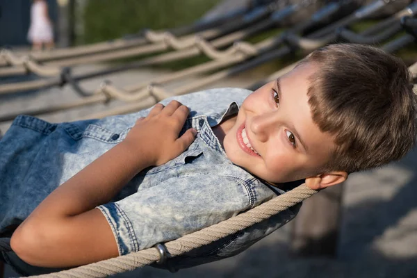 Sarışın çocuk halka açık bir parkta oyun parkında uzanırken kameraya bakıp gülümsüyor.. — Stok fotoğraf