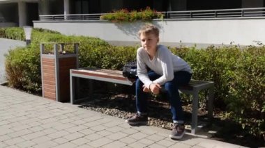 Bir okul çocuğu güneşli bir günde bankta oturur ve etrafına bakar.