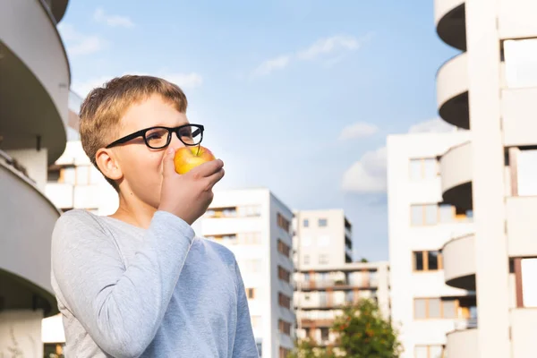 Cute uczennica z okularami jedzenie jabłka przed błękitnym niebem w słoneczny dzień. — Zdjęcie stockowe