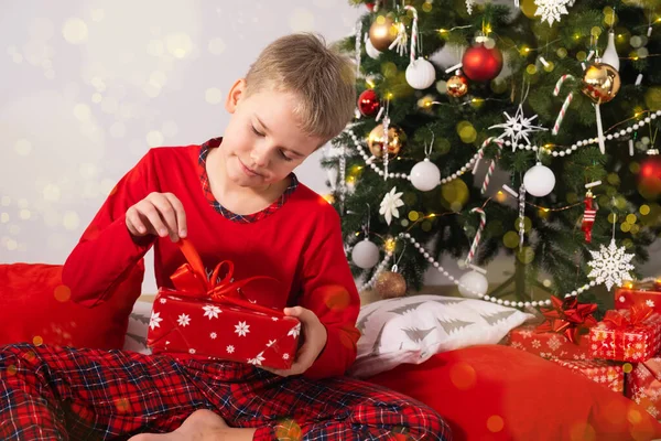 Щасливий хлопчик у червоній піжамі сидить на килимі і відкриває подарунок. Різдвяна ялинка з прикрасами на фоні. Щасливе дитинство . — стокове фото