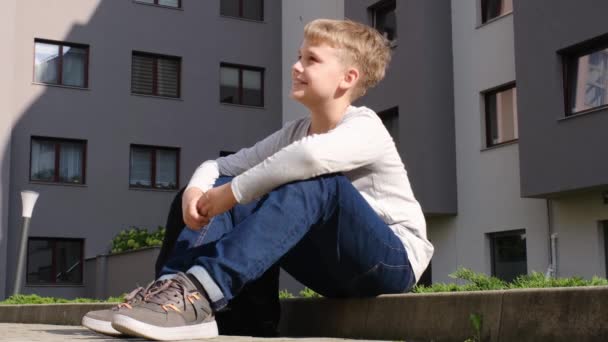 Schoolboy is sitting on curb in a good mood and talking to someone on sunny day — Stock Video