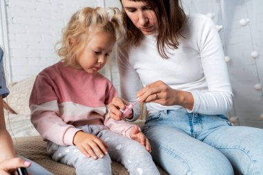 Anne ve kızları yatak odasındaki yatağa oturur ve birbirlerine ojeyle oje sürerler..