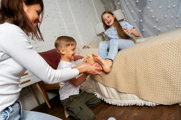 Mama w sypialni na łóżku zabawy łaskocze nogi dzieci, chłopców i dziewcząt. — Zdjęcie stockowe