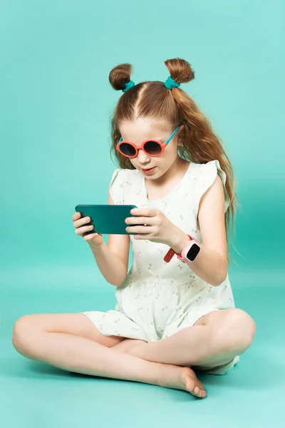 Mobile gaming: a girl in sunglasses sits and plays on a smartphone. Studio portrait on a green background.