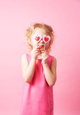 Küçük kız pembe bir arka planda büyük kalp şeklinde lolipoplar tutuyor..