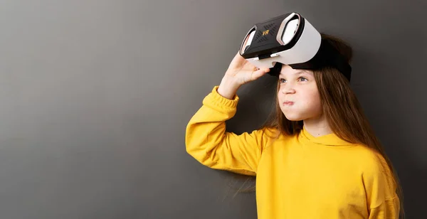 Enthusiastic little girl wearing virtual reality headset modern innovation device isolated on gray studio background. Female kid playing vr game in electronic helmet enjoying futuristic immersion