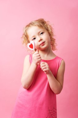 Küçük kız pembe bir arka planda büyük kalp şeklinde lolipoplar tutuyor..
