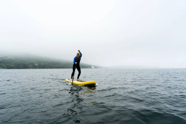 Homem de fato de mergulho azul está remando em uma placa SUP amarela em ondas do mar em dia nublado. — Fotografia de Stock