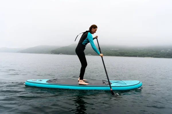 Mulher fitness em pé na prancha de surf desfrutando de supsurf com remo em névoa de água do mar sem fim — Fotografia de Stock