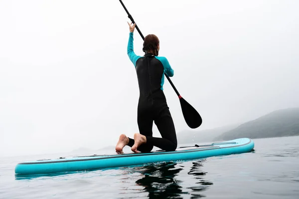 Visão traseira esportista ativa em terno de mergulho flutuando em stand up paddle no cenário do mar de nevoeiro — Fotografia de Stock