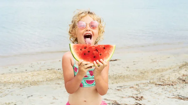Портрет маленькая девочка облизывает свежий кусок арбуза позируя на пляже весело и положительные эмоции — стоковое фото