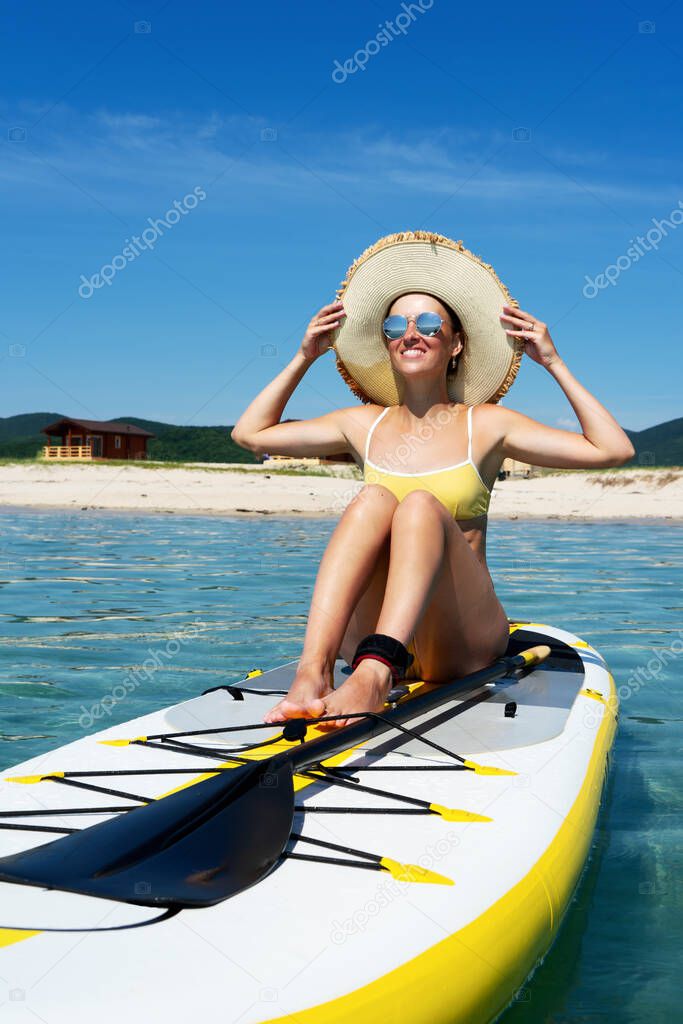 Happy travel woman in straw hat and sunglasses smiling sitting on sup surfboard sunbathing