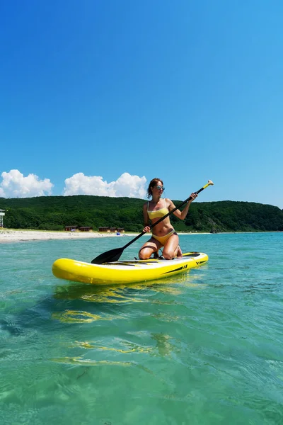 Bikinili bronzlaşmış bir kadın sörf tahtasında oturmuş, yazın keyfini çıkartıyor.