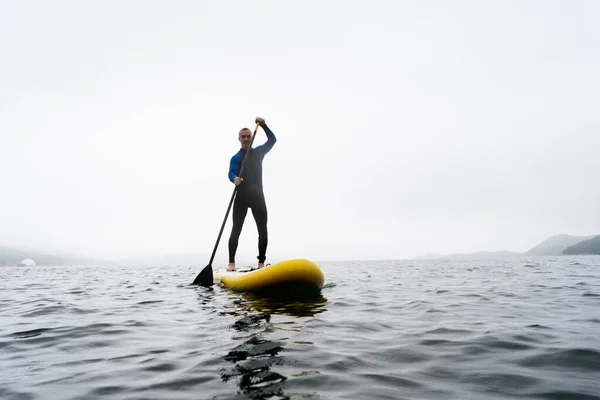 Homem de fato de mergulho azul está remando em uma placa SUP amarela em ondas do mar em dia nublado. — Fotografia de Stock