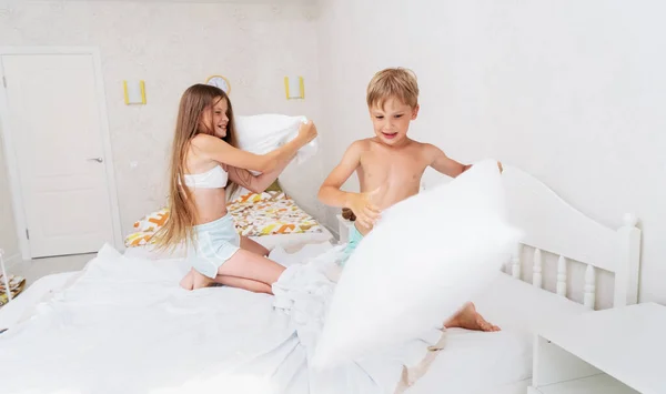 Komik erkek ve kız kardeş yastıkta kavga ediyor. Sabah yatak odasında çocuklar birlikte oynuyorlar. — Stok fotoğraf