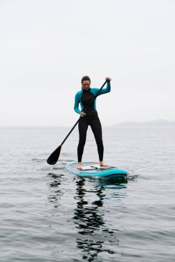 Sörf tahtasında duran sporcu kadın sonsuz deniz sisinde kürekle sörf yapmaktan zevk alıyor.