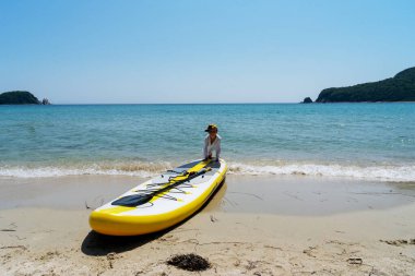 Sahilde sörf tahtası yanında poz veren komik çocuk Shaka hareketi yapıyor.