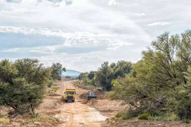VONDELING, Güney Afrika - 21 Nisan 2021: Klaarstroom ve Willowmore arasındaki R407 yolundaki Traka Nehri 'ndeki bir geçitten kum temizleyen bir önyükleyici. Bahşiş kamyonu görünüyor.