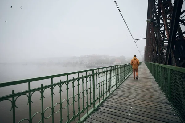 Lonely man walking on bridge against city in mysterious fog. Gloomy weather in Prague, Czech Republic.