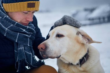 Sıcak giysili bir adam karlı arazide köpeğini okşuyor. Hayvan sahibi ve labrador av köpeği kış manzarasına karşı.. 