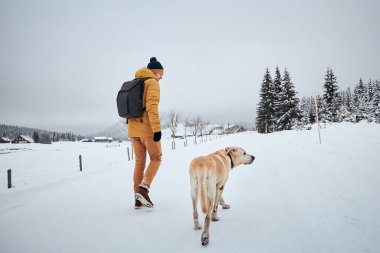 Kışın köpekli genç bir adamın arka görüntüsü. Hayvan sahibi labrador av köpeğiyle eski köye doğru yürüyor. Jizera Dağları, Çek Cumhuriyeti