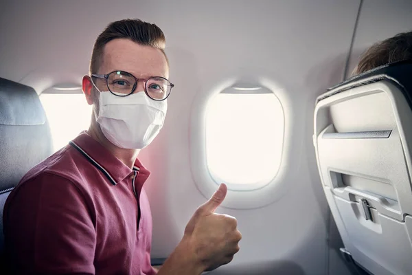 飛行中にフェイスマスクと幸せな観光客の肖像画 若い男が飛行機に親指を立てた テーマは 新しい正常な 個人的な保護とパンデミックで旅行 Covid — ストック写真