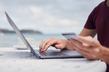 Sahilde denize karşı oturan ve online rezervasyon ya da dizüstü bilgisayarla alışveriş için kredi kartı kullanan bir adam..