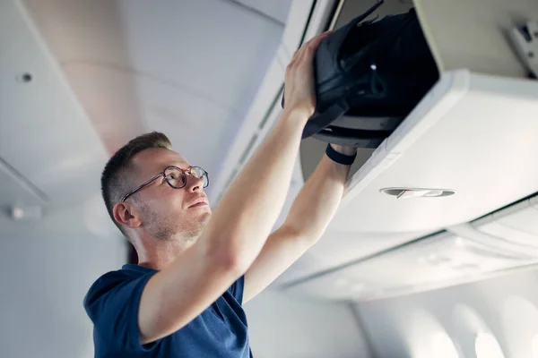 若い男が飛行機で旅行する 飛行機の座席の上のロッカーに手荷物を入れる乗客 — ストック写真