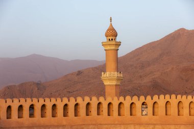 Nizwa Kalesi 'nin duvarları ve El Kala Camii' nin Nizwa, Umman 'da gün ışığıyla aydınlanan dağlara bakan görüntüsü. Tarihi mimari, sıcak ışık ve geleneksel Orta Doğu atmosferi..