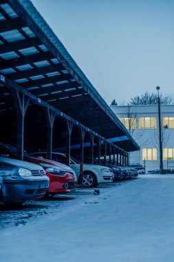 Kar yağışlı bir kış gününde ahşap bir garajın altına park edilmiş bir sıra araba ve arka planda bir bina.