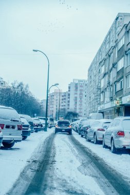 Apartman binaları ve gökyüzünde bir kuş sürüsü olan şehirde, karla kaplı bir caddede arabaları park edip sürüyorlar.