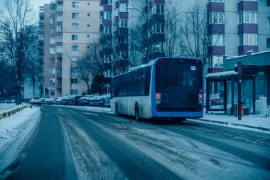 Mavi bir otobüs karla kaplı bir yolda ilerliyor. Apartman binaları caddeyi kaplıyor.